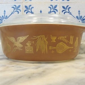 Puede incluir: Una cacerola marrón con tapa de vidrio. La cacerola tiene un diseño dorado de un pájaro, maíz, un mortero y una maja, trigo y una botella.