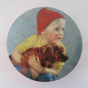 Könnte beinhalten: Eine runde Dose mit blauem Hintergrund, auf der ein kleines Kind mit roter Mütze und gelbem Hemd einen braunen und weißen Hund hält.