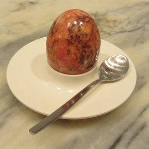 Könnte beinhalten: Ein weißer Keramik-Eierbecher mit einem rot-braunen marmorierten Ei darin. Ein silberner Teelöffel liegt auf einer weißen Untertasse neben dem Eierbecher.