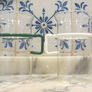 May include: Two clear glass cups with handles. The cup on the left has a green handle and the cup on the right has a white handle.