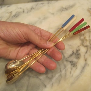 Könnte beinhalten: Ein Satz goldfarbener Löffel mit farbigen Griffen. Die Griffe sind in Blau, Weiß, Rot und Grün bemalt. Die Löffel werden in einer Hand gehalten. Der Hintergrund ist Marmor.