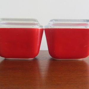 May include: Two red glass storage containers with clear lids. The containers are square shaped and have a white rim.