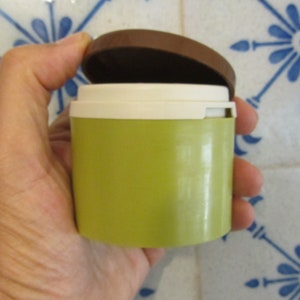 May include: A small, round, green container with a white lid and a brown top. The container is being held in a hand.