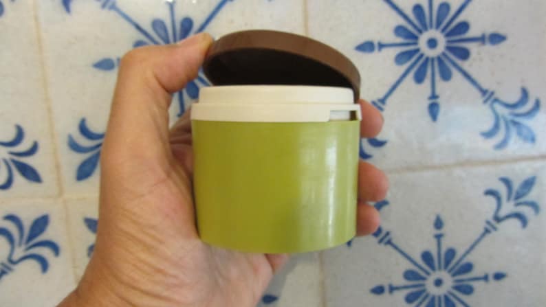 May include: A small, round, green container with a white lid and a brown top. The container is being held in a hand.