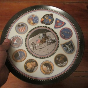 Può includere: Un piatto decorativo rotondo con le missioni Apollo. Il piatto ha un bordo nero con stelle bianche e un'immagine centrale dell'allunaggio. Intorno al centro ci sono toppe circolari con emblemi della missione. Il piatto è tenuto da una persona.