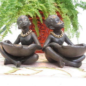 Puede incluir: Dos figuras de piel oscura en posición sentada, cada una sosteniendo un plato en forma de cuenco. Llevan collares de cadena dorada. El fondo incluye una maceta roja con plantas verdes y un mantel estampado.