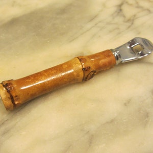 May include: A bamboo bottle opener with a silver metal opener. The handle is made of natural bamboo with a light brown color.