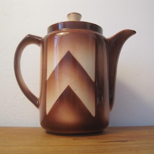 Puede incluir: Cafetera de cerámica marrón con tapa y asa. La cafetera presenta un diseño de chevron en tonos marrones y crema. La cafetera está sobre una superficie de madera.