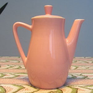 Puede incluir: Una tetera de cerámica rosa con tapa y pico. La tetera está sobre un mantel estampado.