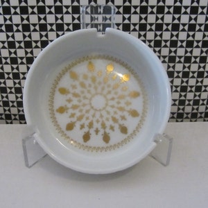 以下が含まれることがあります： 金色の幾何学模様が施された白いセラミック製の灰皿。灰皿は丸型で、中央に小さなへこみがあります。