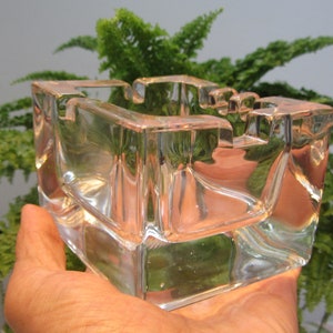 Puede incluir: Un cenicero de vidrio transparente con forma cuadrada y una abertura rectangular en el centro. El cenicero está hecho de vidrio grueso y tiene un aspecto ligeramente esmerilado.