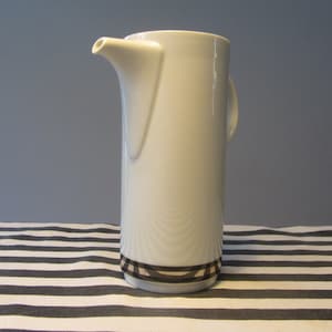 May include: A tall, white ceramic pitcher with a spout and handle. The pitcher has a decorative band of brown and tan squares near the base. The background is a blue-gray wall and a black and white striped tablecloth.
