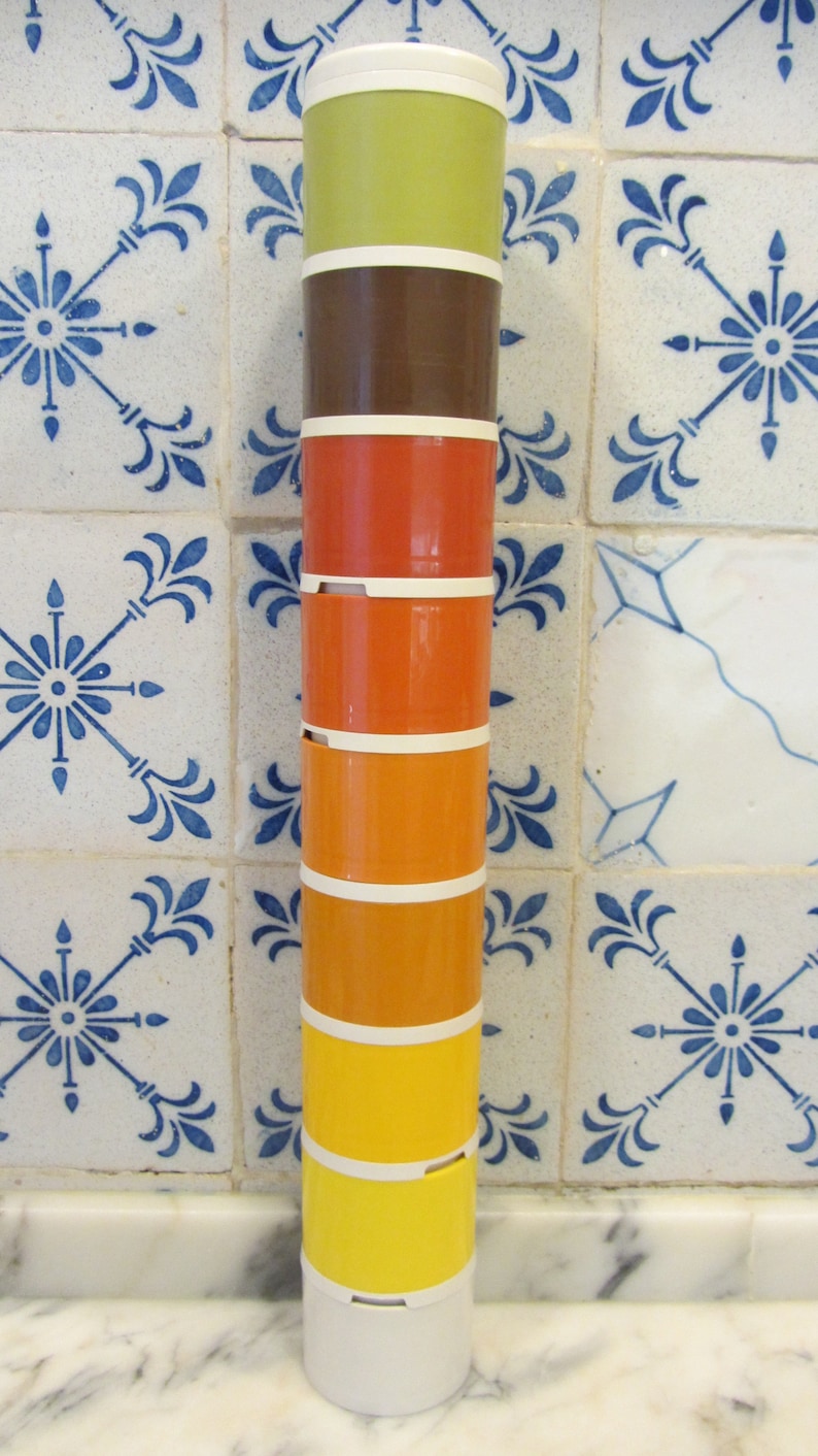 May include: A stackable set of six colourful canisters with a white base. The canisters are stacked in a tower and are made of plastic. The colours of the canisters are green, brown, orange, yellow, and white.