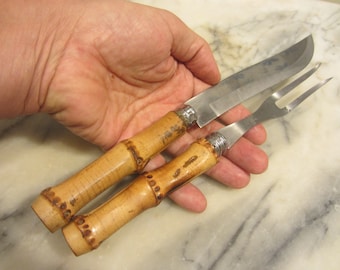 May include: A set of two kitchen utensils with bamboo handles. The knife has a stainless steel blade and the fork has a stainless steel tines. The handles are a light brown color and have a textured surface.
