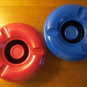 Könnte beinhalten: Zwei runde Aschenbecher, einer rot und einer blau, liegen nebeneinander auf einer Holzoberfläche. Jeder Aschenbecher hat ein kreisförmiges Design mit drei Zigarettenablagen und einer zentralen Vertiefung. Der rote Aschenbecher ist links, der blaue rechts.