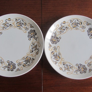 Könnte beinhalten: Zwei weiße Keramikteller mit einem goldenen und schwarzen Blumendesign. Die Teller haben einen weißen Hintergrund mit einem schwarzen und goldenen Blumendesign am Rand.