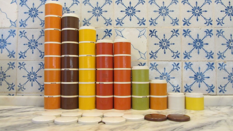 May include: A set of vintage stacking storage containers in various colours, including orange, brown, yellow, green, and white. The containers are arranged in a pyramid shape against a tiled background.