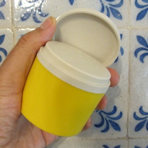 May include: A yellow plastic container with a white lid. The lid is open and the container is being held in a hand.