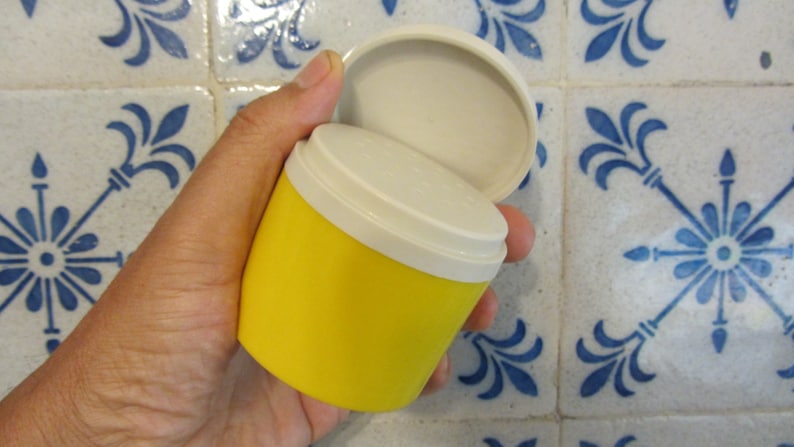 May include: A yellow plastic container with a white lid. The lid is open and the container is being held in a hand.