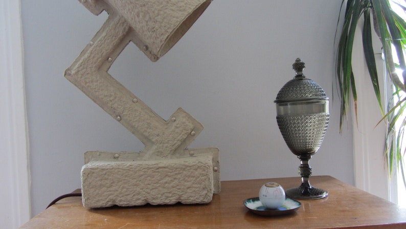 May include: A white, textured lamp with a geometric design sits on a wooden table next to a silver, decorative, glass jar with a lid.