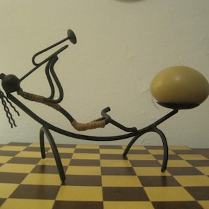 Op de afbeelding: Een zwart metalen beeld van een persoon die op een gebogen basis zit met een ronde, beige kaarsenhouder erbovenop. Het beeld staat op een geruit oppervlak.