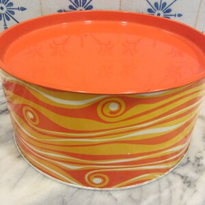 以下が含まれることがあります： オレンジ色の丸い金属製の缶で、お揃いの蓋が付いています。缶の本体は、黄色、オレンジ、白の色合いの波模様で装飾されています。この缶はヴィンテージのようで、状態も良好です。