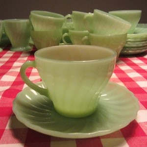 May include: A set of vintage green teacups and saucers. The cups and saucers are made of a translucent, milky green glass. The cups are all different shapes and sizes, and some have a scalloped edge. The saucers are all round and have a scalloped edge.