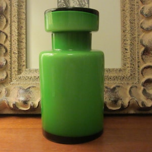 May include: A green glass bottle with a black top. The bottle is cylindrical and has a smooth surface.