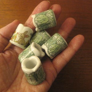 May include: Five small ceramic candle holders with a green and white floral design. The holders feature a variety of designs, including a rabbit, flowers, and leaves.