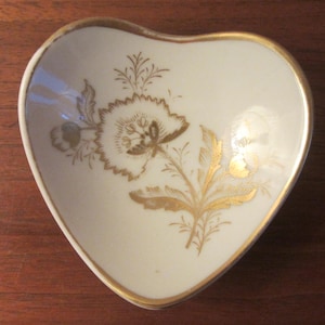 Könnte beinhalten: Herzförmige Schmuckschale mit cremefarbenem Grund und goldenem Blumendesign. Die Schale hat einen goldenen Rand und ein detailliertes botanisches Muster in Gold. Ein dekoratives Stück zur Aufbewahrung kleiner Gegenstände.