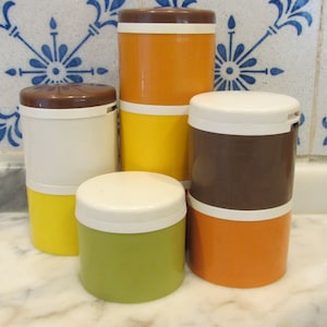 May include: Set of five vintage kitchen canisters with brown, white, orange, and yellow lids. The canisters are stacked on a white marble worktop.