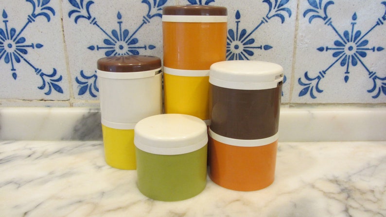 May include: Set of five vintage kitchen canisters with brown, white, orange, and yellow lids. The canisters are stacked on a white marble worktop.