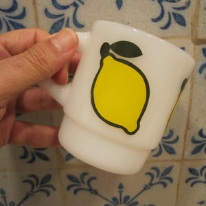 Puede incluir: Taza de cerámica blanca con un contorno negro de un limón con un tallo y una hoja verdes. La taza tiene un asa y está sobre una superficie de azulejos.