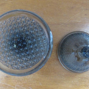 May include: A clear glass jar with a textured lid. The jar has a diamond pattern on the bottom and the lid has a smooth, slightly domed surface.
