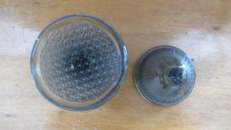 May include: A clear glass jar with a textured lid. The jar has a diamond pattern on the bottom and the lid has a smooth, slightly domed surface.