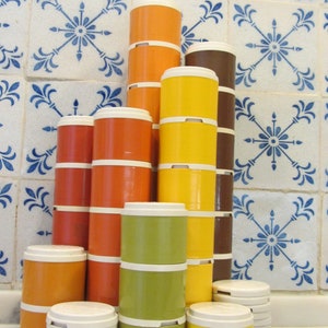 May include: A set of stacked, round, colourful canisters with lids. The canisters are in shades of orange, yellow, green, and brown. The canisters are arranged in a pyramid shape on a white surface.
