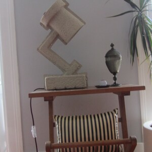 May include: A gray concrete lamp with a unique geometric design sits on a wooden table with a striped pillow and a decorative glass vase. The lamp is plugged in and has a cord running down the side of the table.