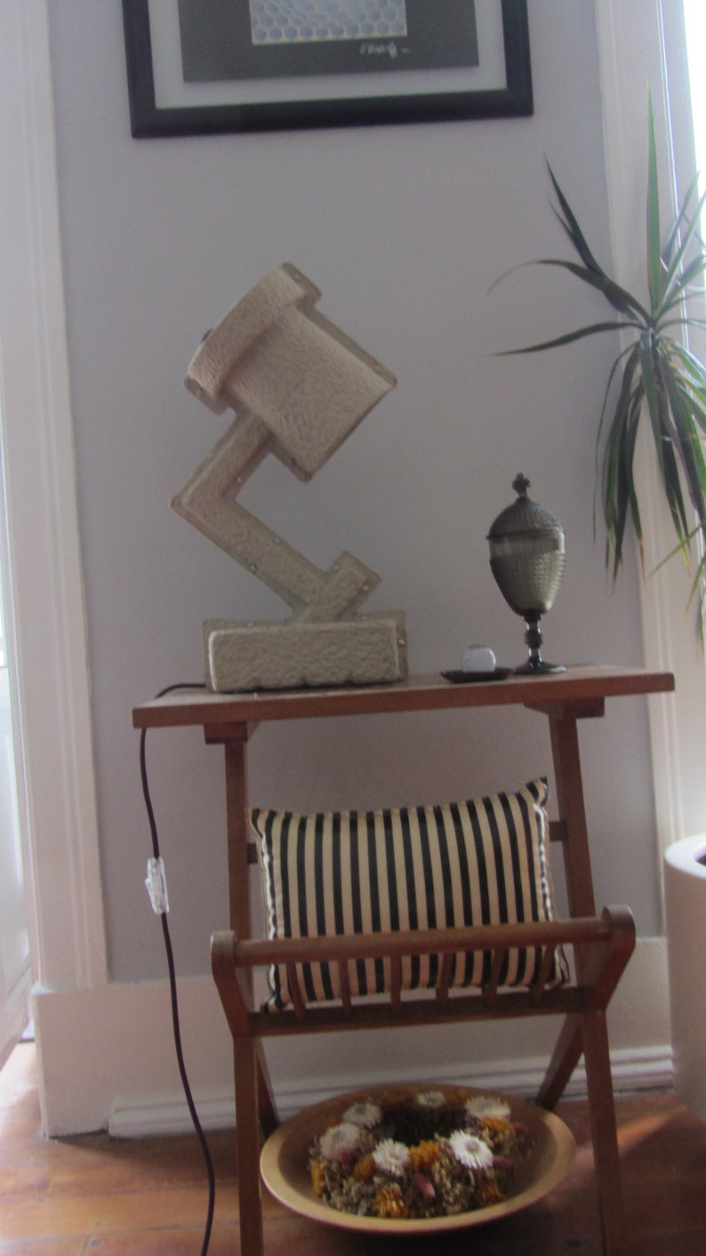 May include: A gray concrete lamp with a unique geometric design sits on a wooden table with a striped pillow and a decorative glass vase. The lamp is plugged in and has a cord running down the side of the table.