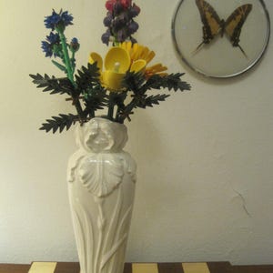 Könnte beinhalten: Eine weiße Keramikvase mit Iris-Design hält einen Strauß bunter Plastikblumen. Die Blumen sind blau, gelb, grün und lila. Die Vase steht auf einer karierten Oberfläche.