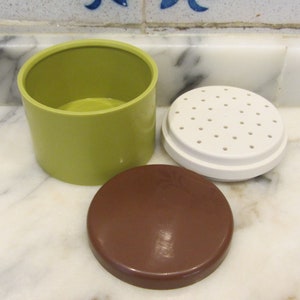 May include: A green plastic container with a white lid with holes and a brown lid. The container is for storing and dispensing spices.