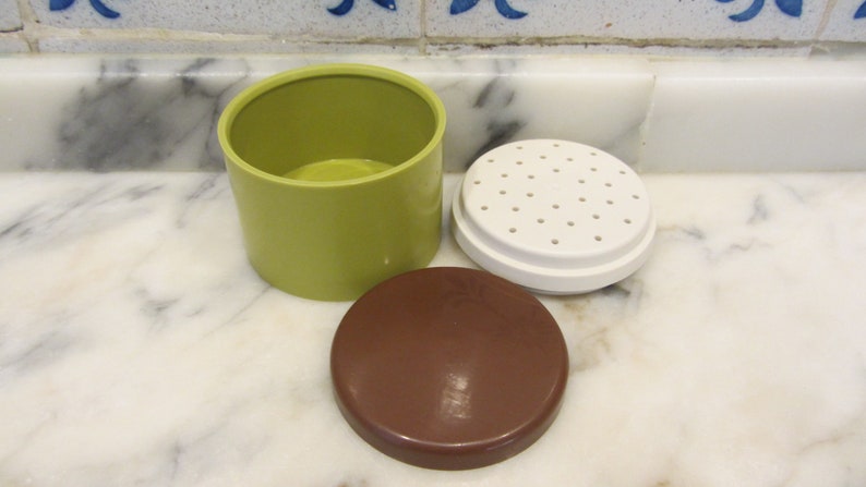 May include: A green plastic container with a white lid with holes and a brown lid. The container is for storing and dispensing spices.