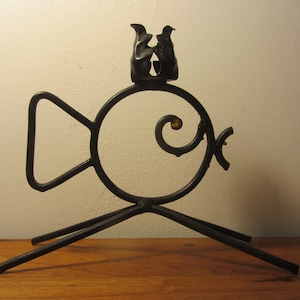 Op de afbeelding: Zwarte metalen kaarsenhouder in de vorm van een vis met een decoratieve swirl aan de zijkant. De houder heeft een kleine, vogelvormige kaarsenhouder bovenop.