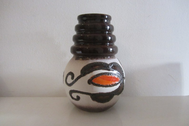 May include: A white ceramic vase with brown and orange geometric designs. The vase has a wide, round body and a tall, cylindrical neck with four stacked rings.