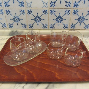 May include: A set of clear glass serving pieces, including small pitchers and bowls, arranged on a rectangular wooden tray. The tray has a reddish-brown finish. The background features a white marble surface and blue and white patterned tiles.