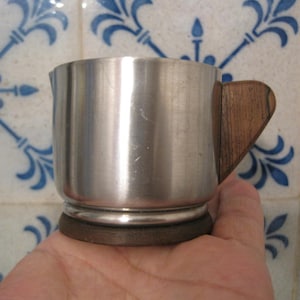 Könnte beinhalten: Eine kleine Kaffeekanne aus gebürstetem Metall mit Holzgriff. Die Kaffeekanne hat einen dunklen Holzsockel und einen Ausguss zum Ausgießen. Der Griff ist hellbraun. Die Kaffeekanne wird in einer Hand gehalten.