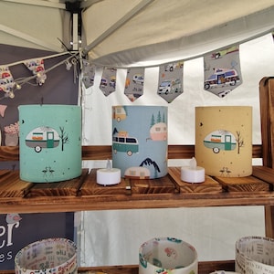 Op de afbeelding: Drie cilindrische kaarshouders in de kleuren turquoise, blauw en geel met een camper-ontwerp. De kaarshouders staan op een houten plank met twee witte theelichtjes.