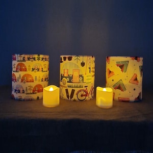 Könnte beinhalten: Drei zylindrische Kerzenhalter mit Camping-Motiv. Jeder Halter hat ein anderes Design mit einem Wohnwagen, einem Fahrrad und einem Zelt. Die Kerzenhalter sind mit kleinen, runden, weißen LED-Kerzen beleuchtet.