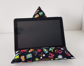 Puede incluir: Una tableta negra apoyada en una almohada negra y blanca con un estampado de temática de camping. La almohada tiene una variedad de iconos de camping coloridos, incluyendo una tienda de campaña, una linterna, una fogata y una brújula.