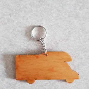 Puede incluir: Llavero de madera con forma de autocaravana con un anillo y una cadena de metal plateado. El llavero es de color marr&oacute;n claro y tiene un acabado liso.