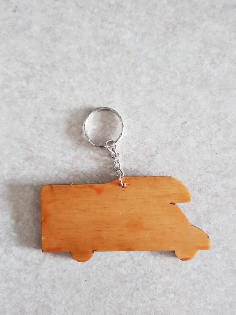 Puede incluir: Llavero de madera con forma de autocaravana con un anillo y una cadena de metal plateado. El llavero es de color marr&oacute;n claro y tiene un acabado liso.